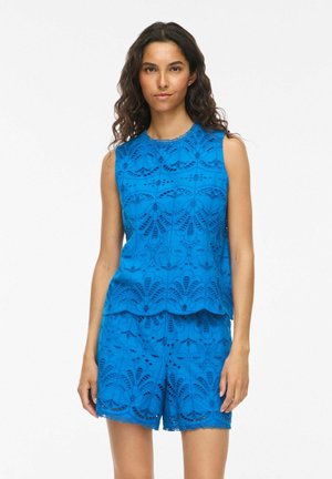 Femme portant un haut en dentelle bleu vif sans manches et un short assorti avec des motifs floraux découpés, debout devant un fond blanc uni.