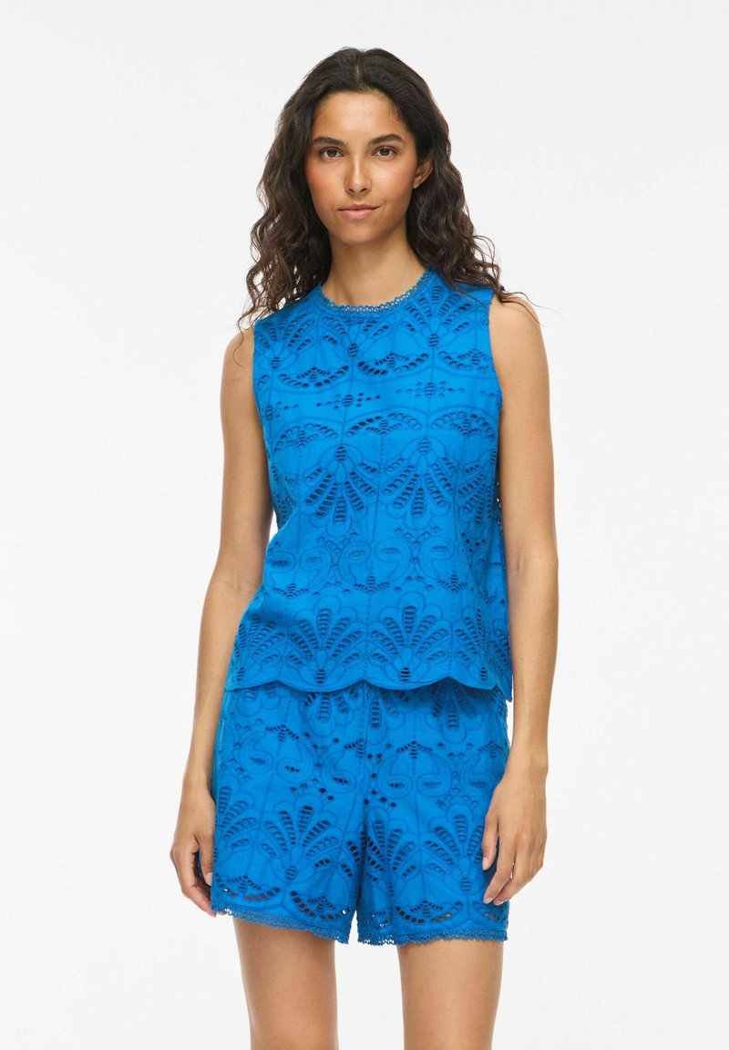 Femme portant un haut en dentelle bleu vif sans manches et un short assorti avec des motifs floraux découpés, debout devant un fond blanc uni.