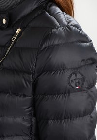 Svart puffjacka med quiltad textur, med en guldfärgad dragkedja och broderad logotyp på ärmen. Mjuk, glänsande tyg.