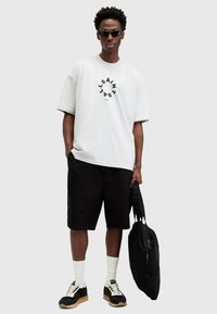 Hvit oversized t-skjorte med svart sirkulært logo, svarte shorts, svarte joggesko med kremfargede detaljer, hvite sokker og en svart tote-bag.
