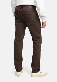 Brązowe dżinsy z slim fit, pięcioma kieszeniami i markowym paskiem, w połączeniu z białymi trampkami na neutralnym tle.