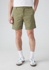 Oliivinvihreät puuvillashortsit suoran leikkauksen, etutaskujen ja raidan helman kanssa. Yhdistetty valkoisten sukkien ja tennareiden kanssa rentoon ilmeeseen.