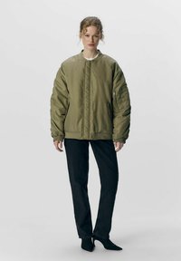 Olivengrøn bomberjakke med lige snit, ribbede manchetter og lynlås-lommer. Båret med sorte lige ben jeans og spidse sko.