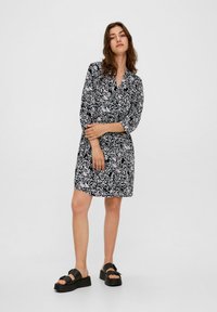 Schwarz-weiß gemustertes Kleid mit einem kragenlosen Halsauschnitt, Dreiviertelärmeln und tailliertem Schnitt. Kombiniert mit schwarzen Plateau-Sandalen.