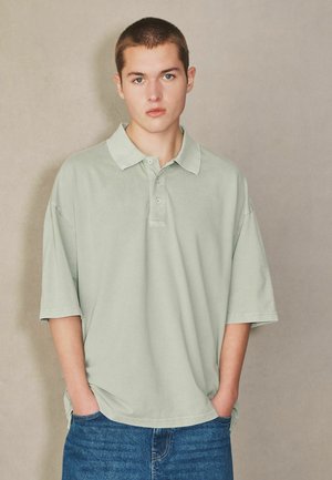 Junger Mann mit kurzem Haar, der ein hellgrünes, übergroßes Poloshirt und blaue Jeans trägt, steht mit den Händen in den Taschen vor neutralem Hintergrund.