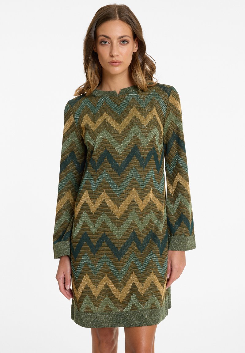 Woman wearing a knee-length, long-sleeve dress with a green, gold, and brown zigzag pattern, standing against a plain white background.