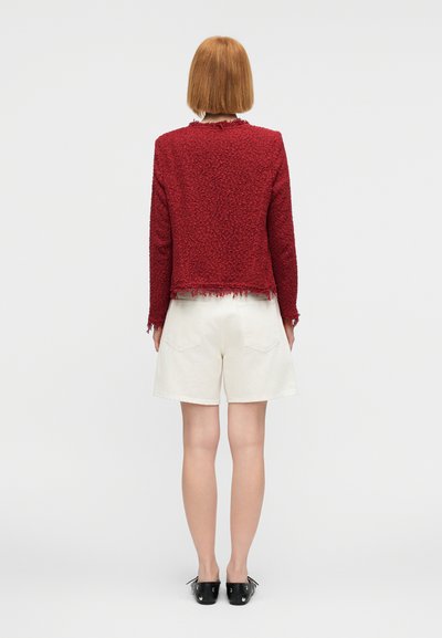 Κόκκινο υφασμάτινο μπουφάν με ξεφτισμένο τελείωμα, συνδυασμένο με μπεζ σορτς. Η εμφάνιση ολοκληρώνεται με μαύρα παπούτσια, από την πίσω όψη.