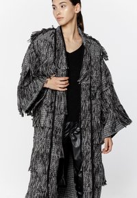 Frędzlowany czarnobiały teksturowany płaszcz z otwartym przodem, szerokimi rękawami i pionowymi czarnymi akcentami. Nosi się go na dopasowanej czarnej bluzce.