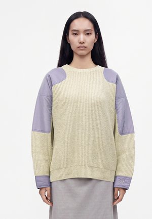 Kvinde med langt sort hår iført en beige striktrøje med lyse lilla stoflapper på skuldre, ærmer og manchetter, stående mod en hvid baggrund.