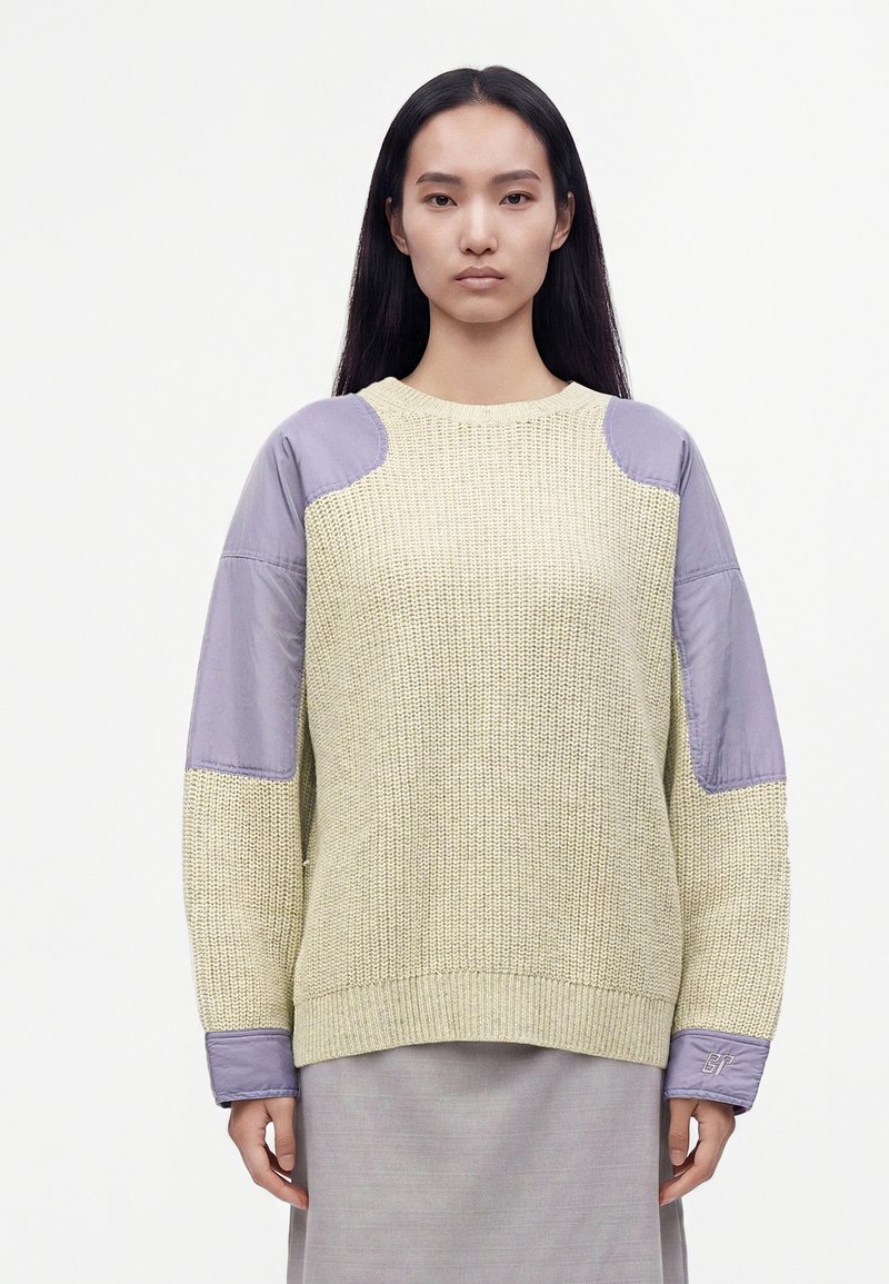 Femme aux longs cheveux noirs portant un pull en maille beige avec des patchs en tissu violet clair sur les épaules, les manches et les poignets, debout sur un fond blanc.