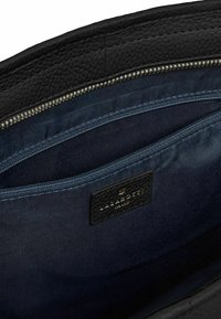 Interno di una borsa a tracolla in pelle nera con una fodera in tessuto blu scuro, una tasca con zip e un'etichetta di marca testurizzata.