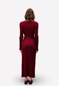 Lange burgundy ribjurk met een getailleerd silhouet en lange mouwen, voorzien van een ceintuurdetail dat in een strik aan de taille is gebonden.