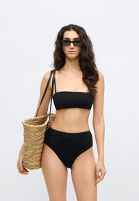 Μαύρο μπικίνι με στράπλες τοπ και σλιπ ψηλόμεσο, συνοδευόμενο από πλεκτή τσάντα από ψάθα με σκούρα δερμάτινα λουριά.