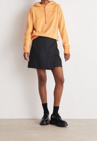 Persona che indossa un felpa pesca con logo "BOSS", una gonna nera avvolgente con orlo sfilacciato, calze nere e scarpe platform nere chunky, in piedi su un pavimento beige.