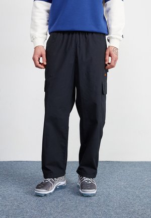 Mand iført sorte cargobukser, grå og hvide sneakers og en blå og hvid langærmet sweatshirt, stående på blåt tæppe.