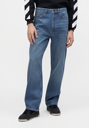 Personne portant un jean bleu à jambes larges associé à des baskets noires et à un haut à manches longues noir avec des rayures diagonales blanches sur les manches.