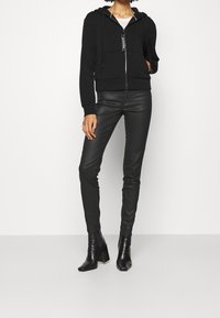 Sudadera con cremallera negra de corte holgado, capucha con cordones y bolsillos frontales. Combinada con pantalones de cuero ajustados negros y botas de tobillo.