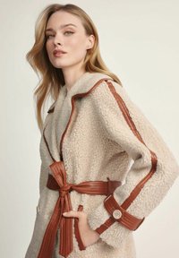 Beżowy płaszcz z faux shearling z brązowymi skórzanymi akcentami, z paskiem w talii i guziczkami na mankietach, prezentujący teksturowaną powierzchnię.