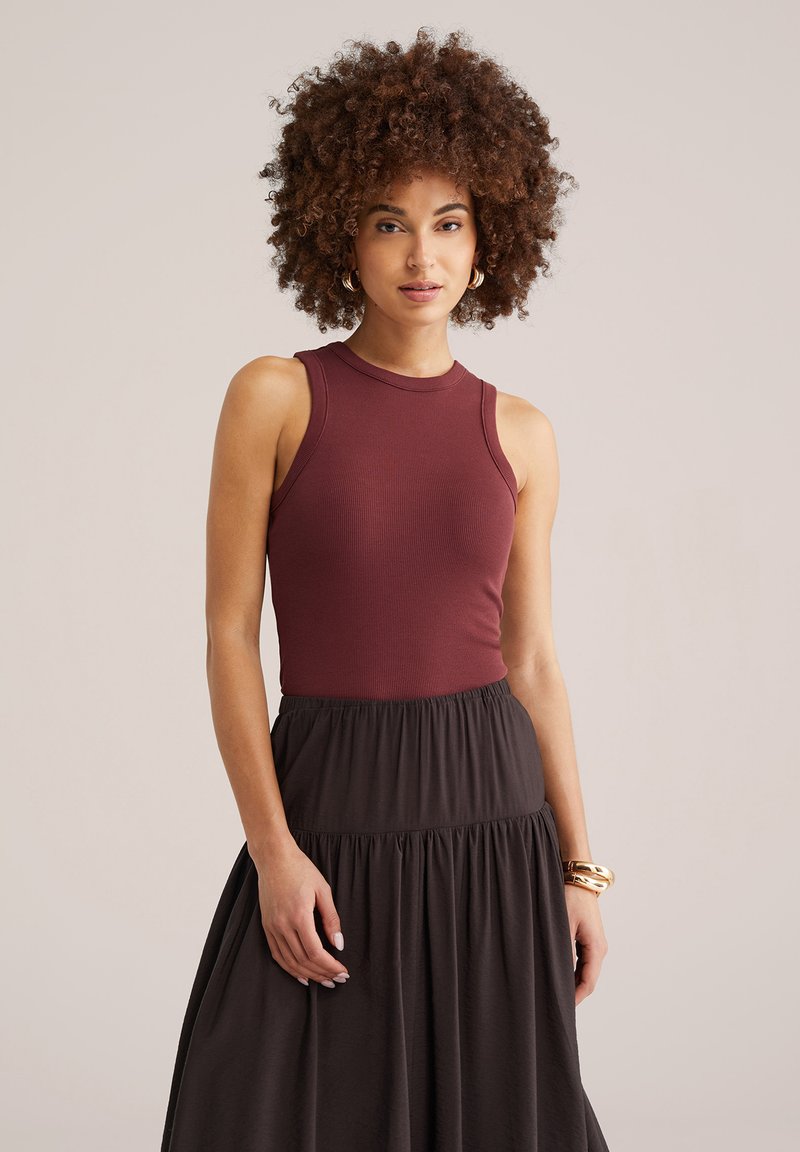 Femeie cu păr creț, purtând un top fără mâneci bordo și o fustă neagră cu talie înaltă, accesorizată cu cercei de aur rotunzi și brățări.