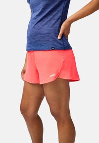 Jasnoróżowe spodenki sportowe z zaokrąglonym dołem, wykonane z lekkiego materiału, z małym logo na lewym udzie, w zestawieniu z niebieską strukturą topu.