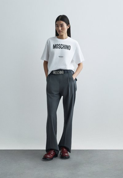 Žena stojí oblečená v bielej tričku Moschino, tmavošedých širokých nohaviciach s opaskom Moschino a tmavočervených topánkach na pozadí bez detailov v šedej farbe.