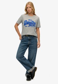 Szara bawełniana koszulka z niebieskim napisem "SuperDry", zestawiona z niebieskimi dżinsami o wysokim stanie i luźnym kroju oraz czarnym obuwiem.