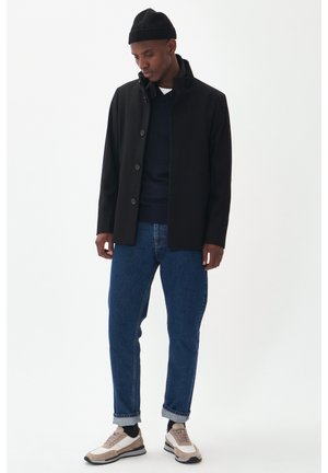 Black buttoned coat with a high collar, worn over a navy sweater, paired with blue jeans and beige sneakers with white accents.