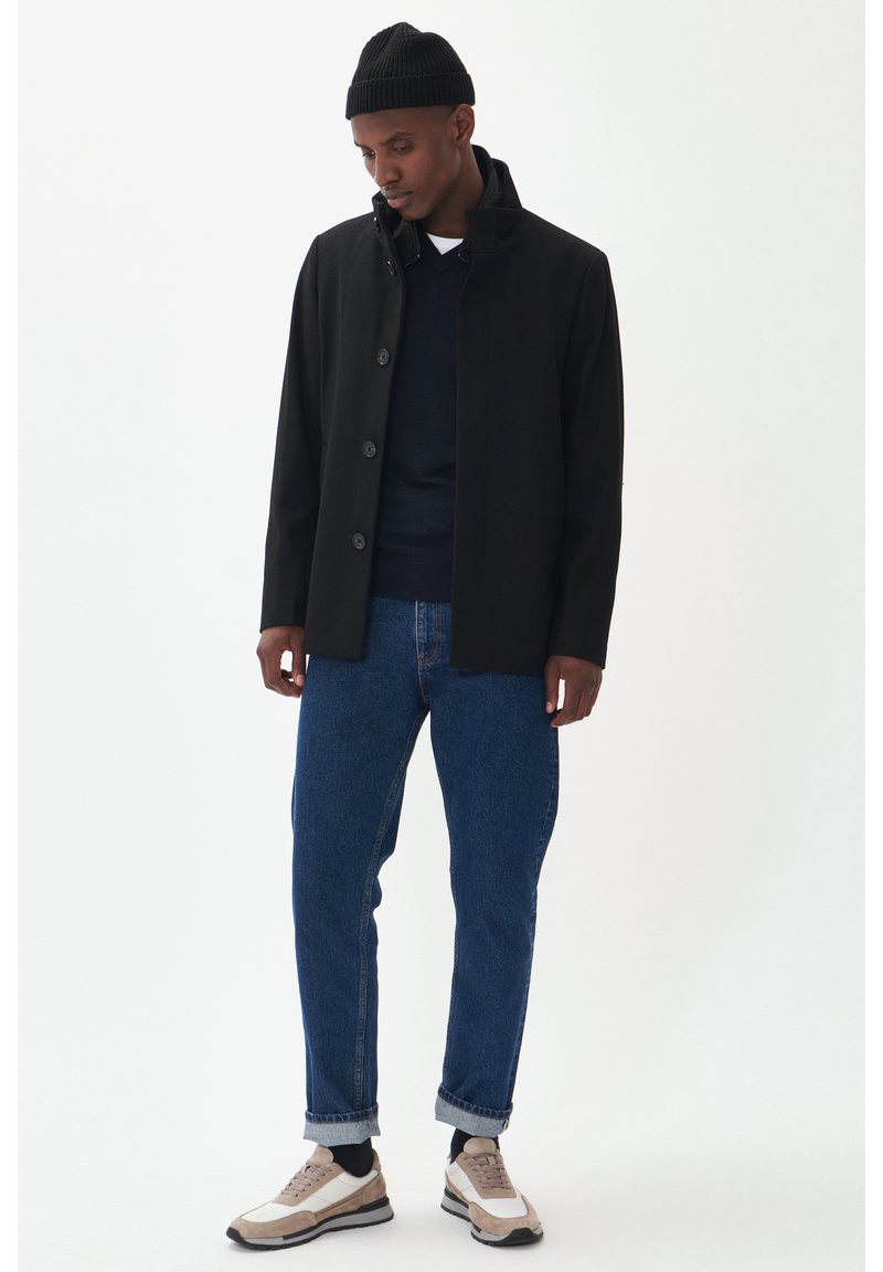 Cappotto nero con bottoni e colletto alto, indossato sopra un maglione blu navy, abbinato a jeans blu e sneakers beige con dettagli bianchi.