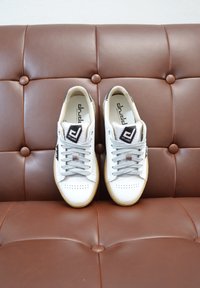 White sneakers with gray laces, perforated toe, and a tan rubber sole. Brand logo on the tongue and side. Positioned on a brown tufted leather surface.