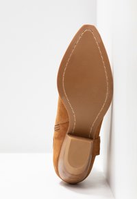 Bruine suède enkellaars op een wit oppervlak, met een gladde zool met gestikte rand en blokhak.