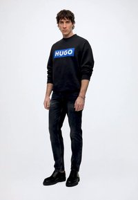Mann trägt schwarzen Pullover mit blauem "HUGO"-Logo, schwarze Jeans und schwarze Schuhe, steht vor schlichtem weißen Hintergrund.