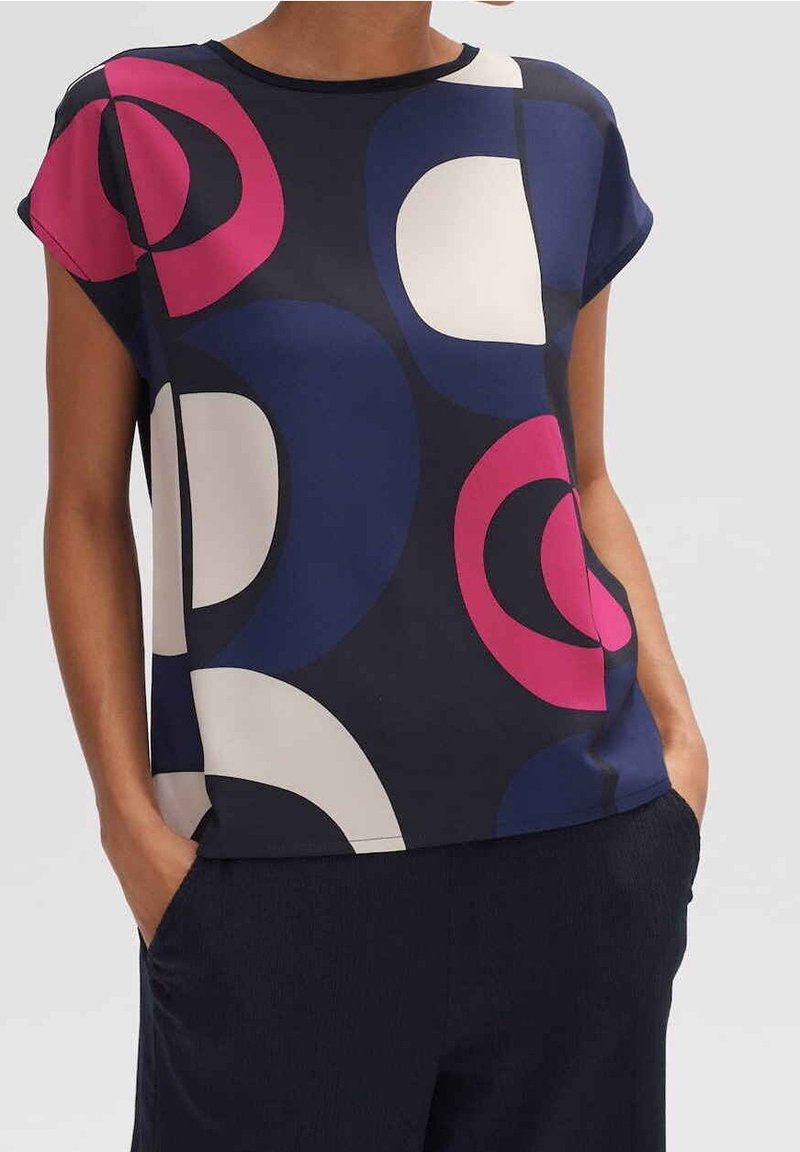 Femme portant un chemisier à manches courtes avec de grands motifs circulaires abstraits en bleu marine, blanc et rose, associé à un pantalon foncé et les mains dans les poches.