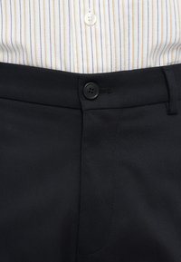 Pantaloni negri cu talie cu nasturi, purtați sub o cămașă albă cu dungi verticale în nuanțe de portocaliu și albastru.