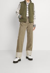 Chaleco acolchado verde sobre una camiseta de tie-dye, combinado con pantalones anchos beige con un dobladillo deshilachado y zapatillas blancas gruesas con cordones negros.