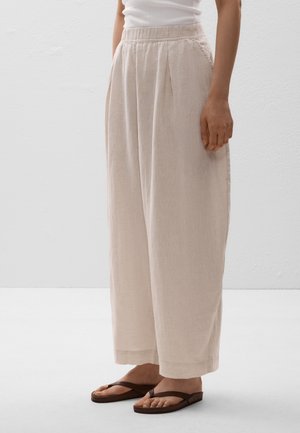 Light beige, loose-fitting trousers with pleats, an elastic waistband, and a soft texture. Paired with dark brown flip-flops.
