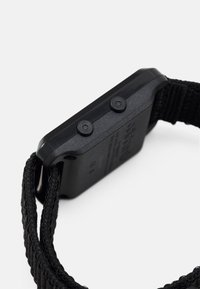 Orologio digitale nero con cassa texturizzata, due pulsanti sul lato e un cinturino in nylon intrecciato. Il retro presenta un testo inciso.
