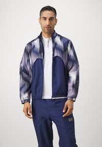 Navy blue jacket with geometric pattern in shades of purple and grey, full zip, elastic cuffs, and logo on the collar and lower hem.