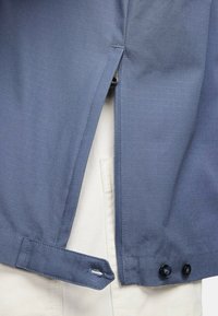 Chaqueta azul oscura con sutiles rayas, que presenta un cierre lateral, puños abotonados y una camiseta clara sólida visible debajo.