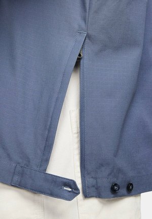Giacca blu scuro con sottili righe, dotata di una cerniera laterale, polsini con bottone e una maglietta leggera di colore chiaro visibile sotto.
