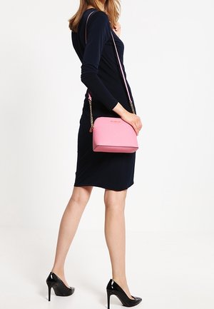 Donna che indossa un vestito nero aderente e tacchi alti neri, mentre porta una piccola borsa a spalla rosa con una tracolla dorata.