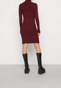 Bordeauxrot geripptes Strickkleid mit langen Ärmeln, hohem Kragen und strukturierten Streifen im oberen Bereich; kombiniert mit knielangen schwarzen Lederstiefeln.