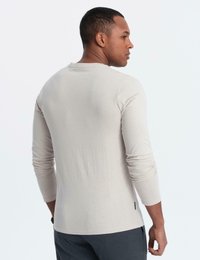 Man i en ljus beige långärmad skjorta och mörka byxor, stående och vänd bort med huvudet vridet åt höger.