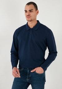 Polo de manga larga en azul marino con cuello, que presenta una tapeta de tres botones. Textura suave, diseño ajustado, combinado con jeans azules.