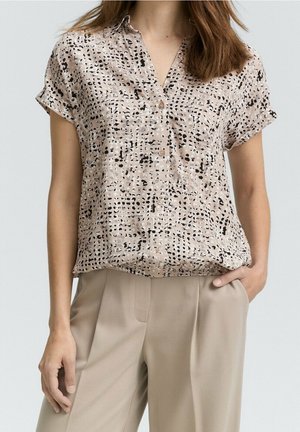 Femme portant un chemisier beige à manches courtes avec des taches noires et un pantalon beige clair, une main dans la poche et les cheveux lâchés.