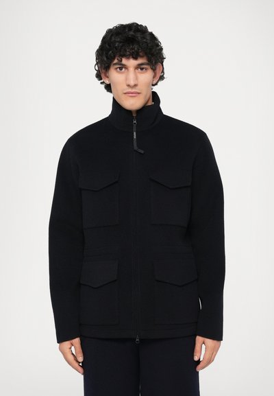 Giacca in fleece nera con colletto alto, zip frontale completa e quattro tasche anteriori. Presenta una superficie testurizzata e una vestibilità sartoriale.