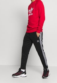 Červená mikina s bílým logem Adidas, černé teplákové kalhoty s bílými pruhy a černé tenisky s růžovými akcenty.