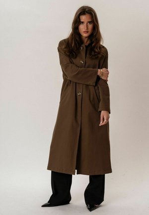 Kobieta z długimi brązowymi włosami, ubrana w brązowy długi płaszcz zapinany na guziki, czarne szerokie spodnie oraz czarne buty na szpic na jednolitym tle.