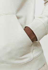 Lys beige sweatshirt med ribbede manchetter og en kængurulomme; blød stofstruktur; kontrasterende brun arm synlig i lommeområdet.