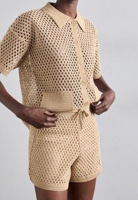 Beige hæklet sæt med kortærmet knap-op top og matchende shorts, begge med diamantnetmønster og snøre i taljen.