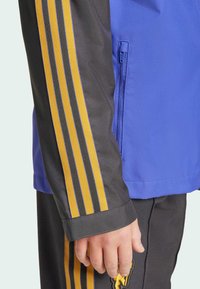 Plava jakna s crnim rukavima ima tri vertikalne žute pruge. Uključuje bočnu džep s patentnim zatvaračem i kontrastne manžete.